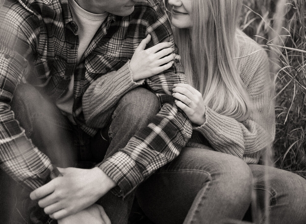 A guy in a flannel and a girl in a sweater are sitting next to each other in the grass and snuggling.