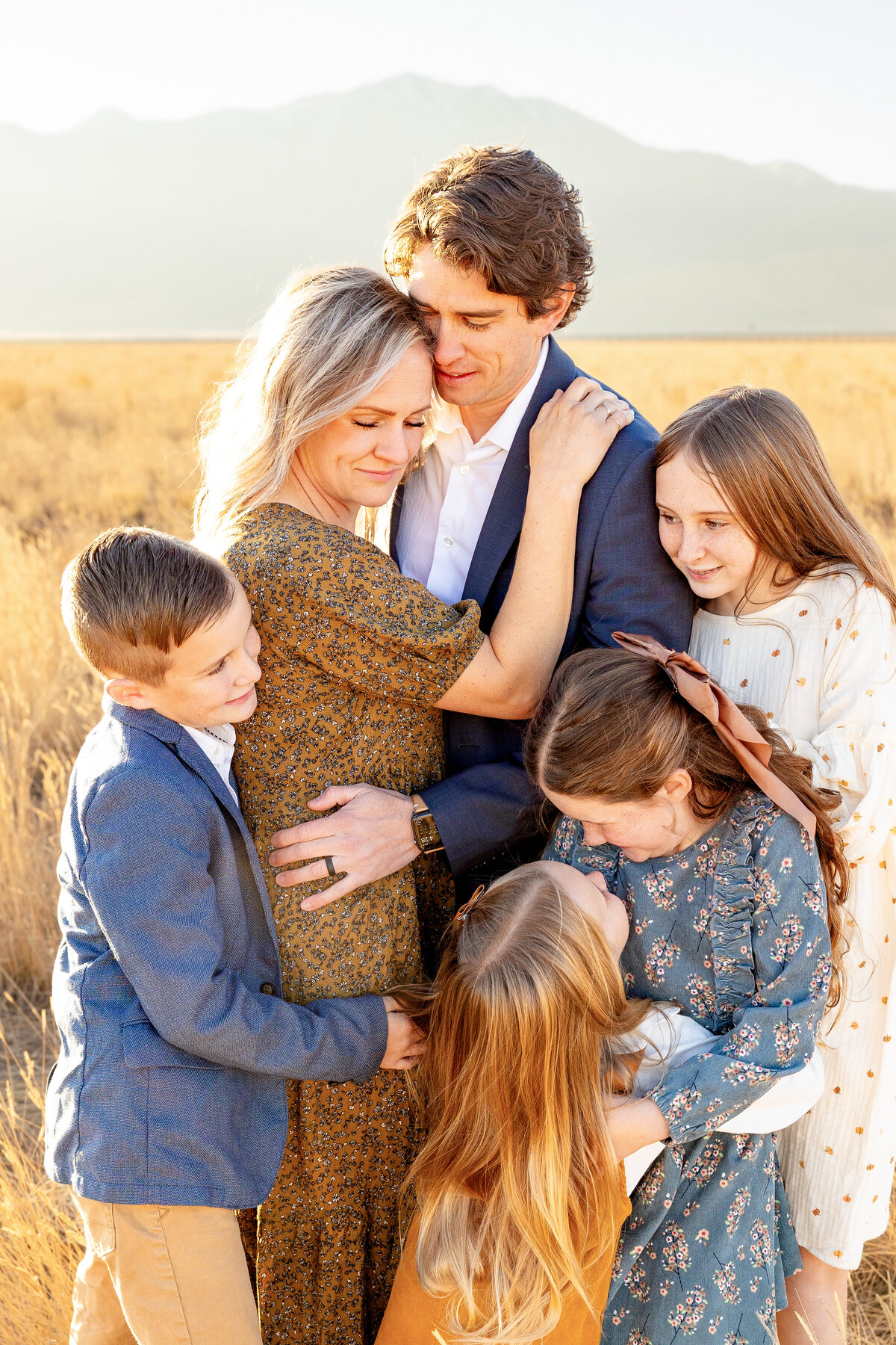 A family of six shares a warm embrace in a golden field, bathed in soft sunset light, with mountains in the distance.