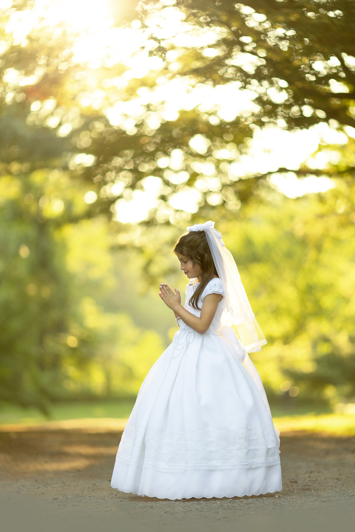 A young girl in a communion dress prays while standing in a park path at sunset