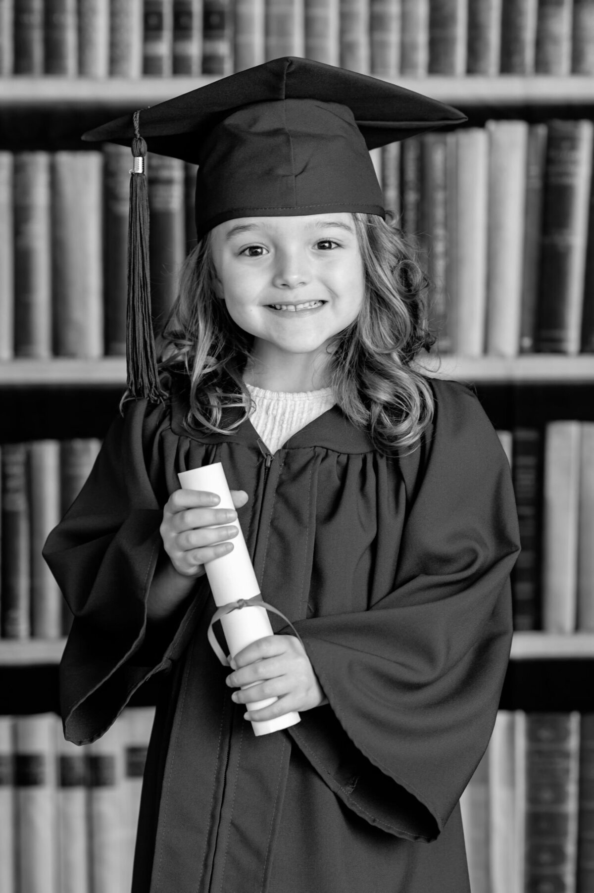The image shows a young girl with curly hair wearing a graduation cap and gown. She is smiling and holding a rolled-up diploma tied with a ribbon. The background consists of shelves filled with books, suggesting a library or academic setting. The girl is centered in the image and appears happy.