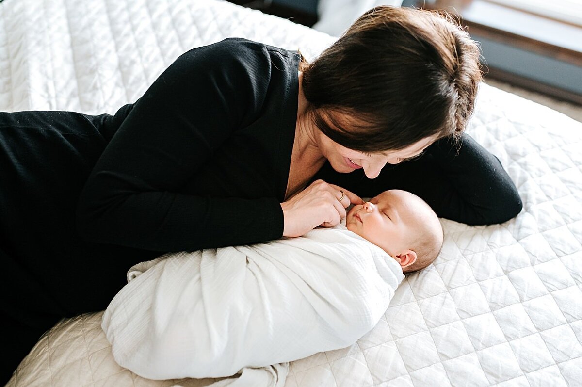 mother lying on bed next to newborn swaddled in white blanket