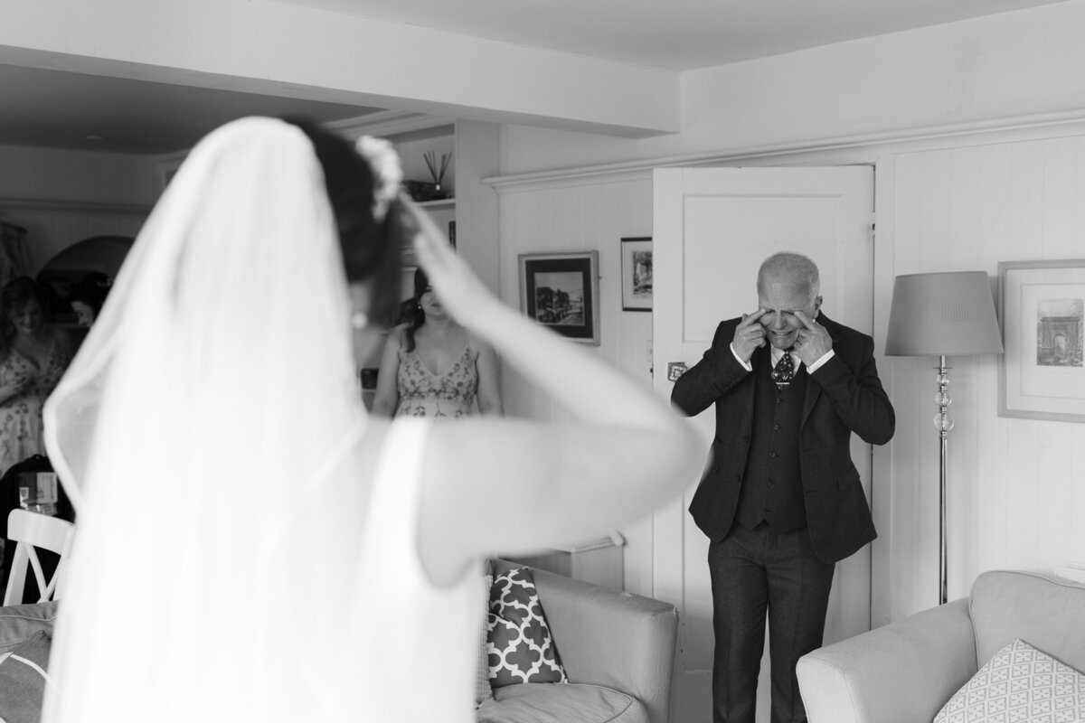 A bride, with her back to the camera, stands in a living room. Her father is visibly emotional and wiping tears from his eyes