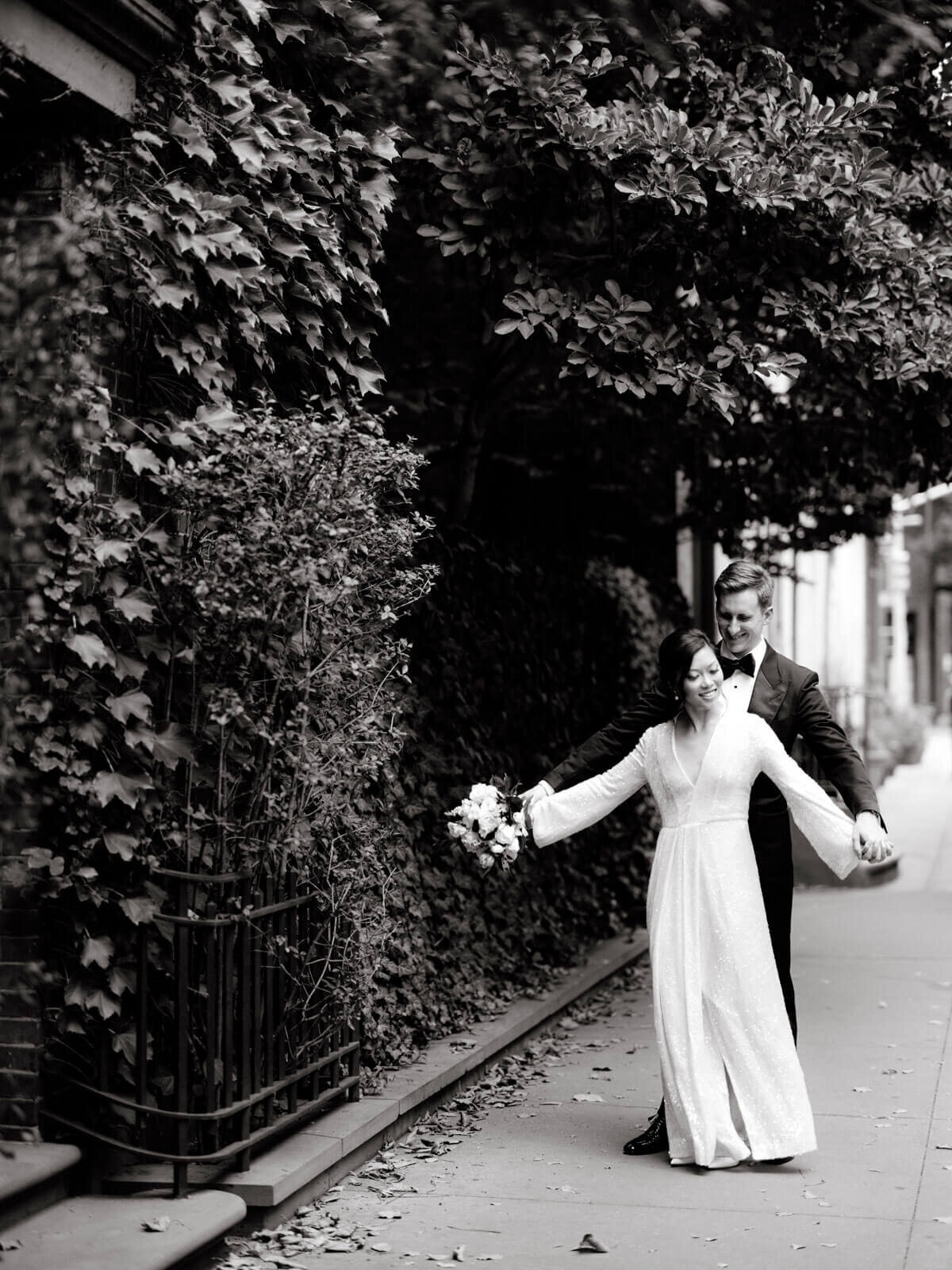 The groom is romantically lifting the bride's hands from behind, along a sidewalk in New York City. Image by Jenny Fu Studio