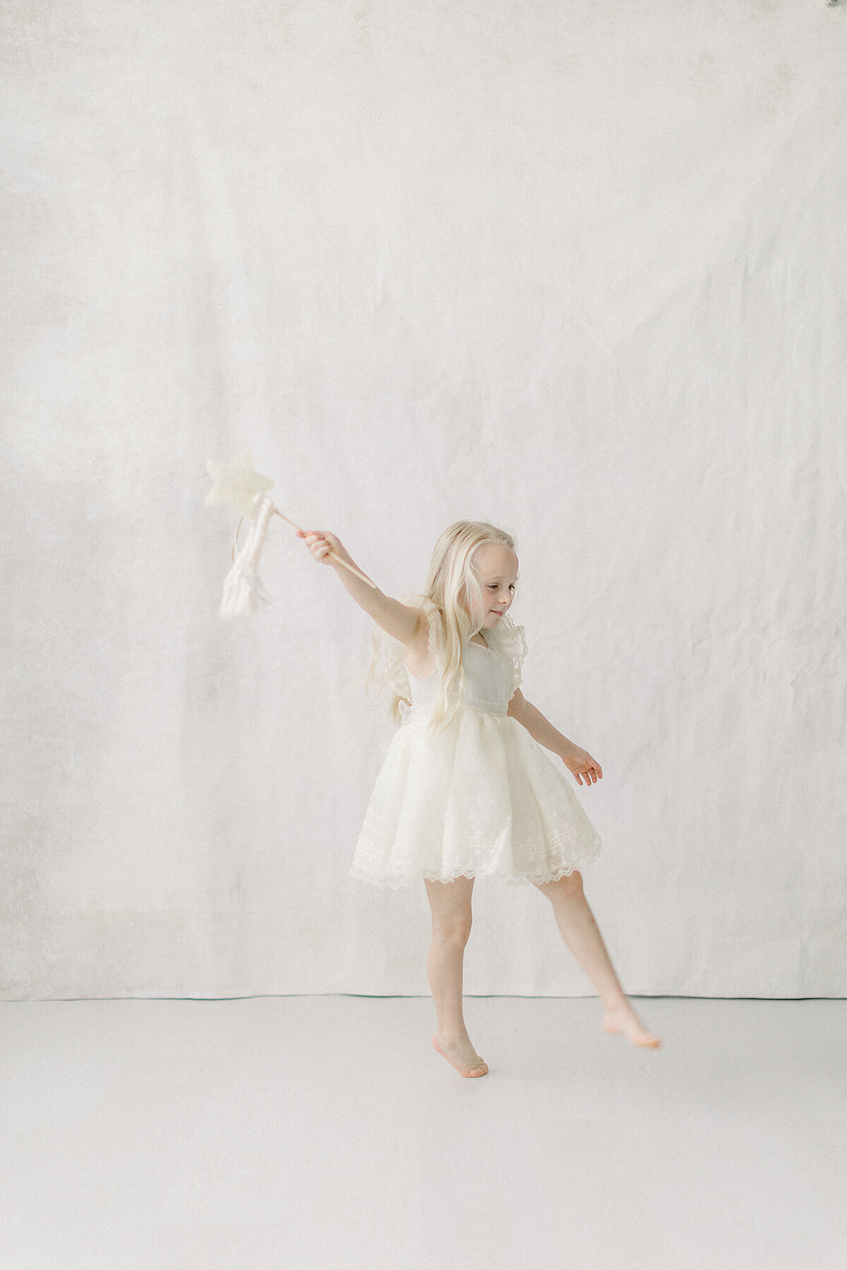 A beautiful ligth filled portrait of a girl dressed in a cream birthday dress dancing around a photography studio in Dallas holding a star wand with ribbon draping down from it.