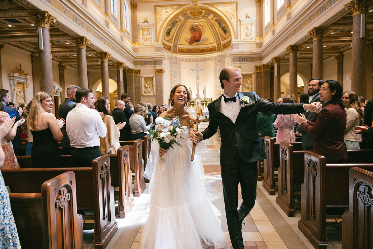 Bride dressed in white and groom in a black tux laugh while walking down the aisle after their wedding ceremony. 