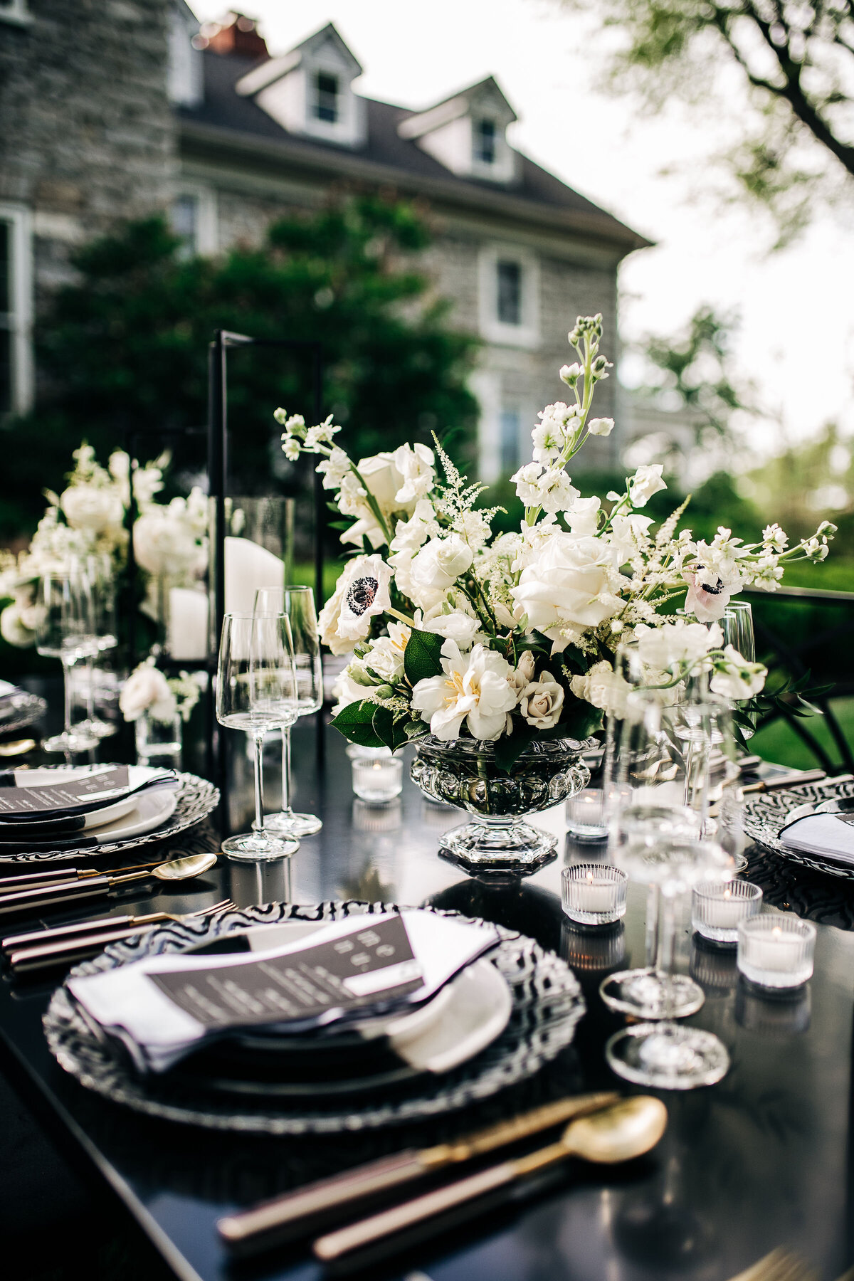 Sophisticated outdoor tablescape featuring white flower arrangements, black accents, and gold flatware on a chic black table setting.