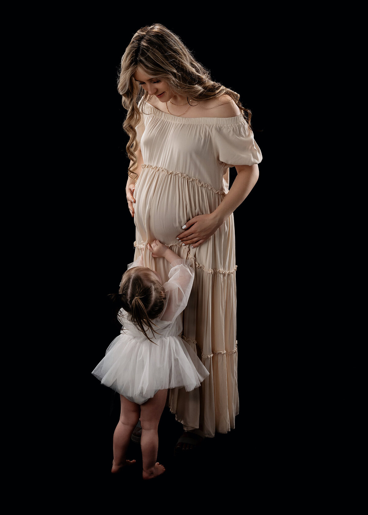A baby girl in a white dress reaches up to mom's pregnant bump while standing in a studio of an Iowa maternity photographer