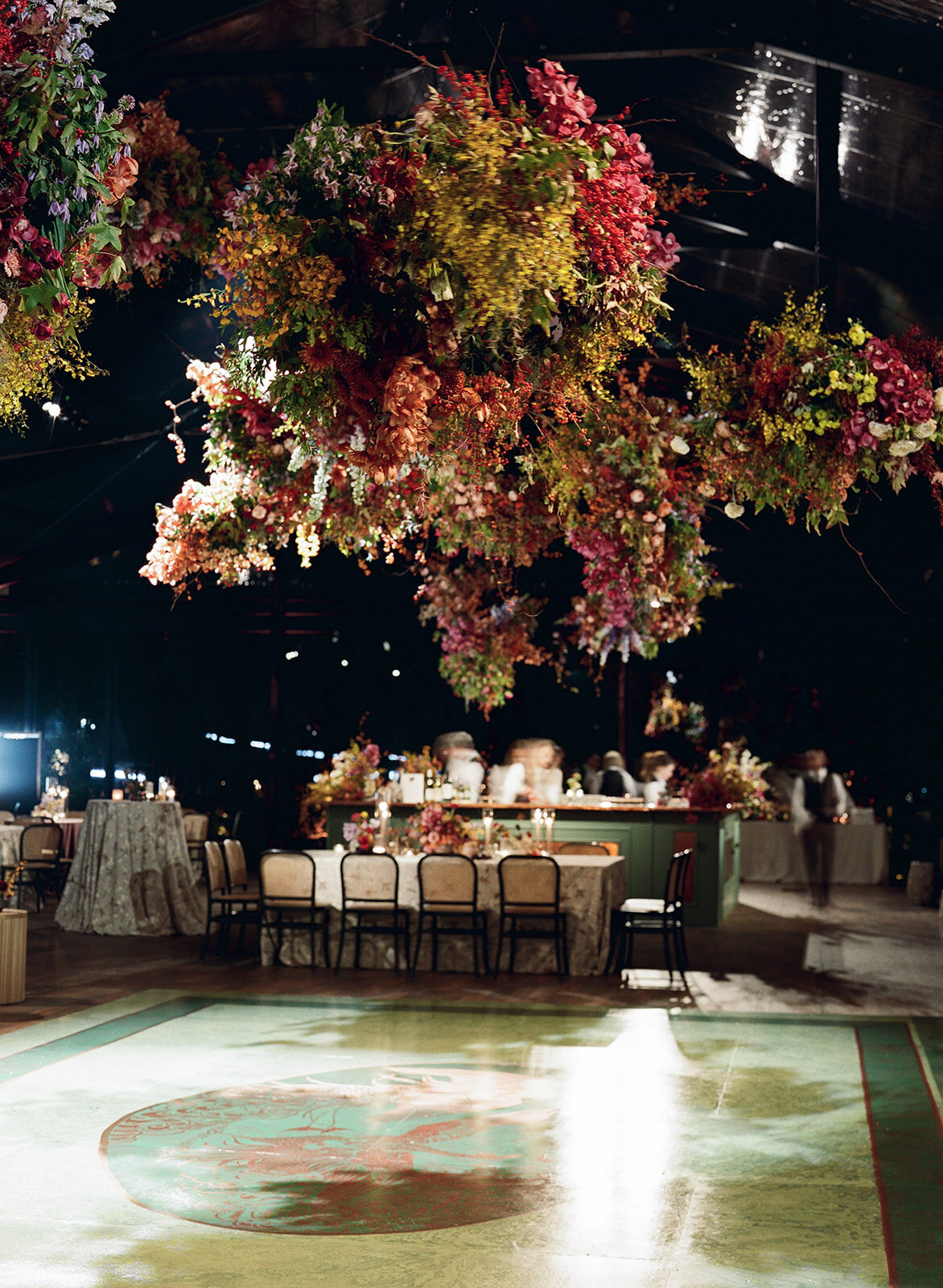 Interacting and sculptural floral clouds hang over the dance floor and bars of this fall wedding reception in Tennessee. Clear top tent build out for fall wedding reception in Tennessee hills.  Product clustered floral clouds in a tasteful fall color palette of copper, ruby, gold, burgundy, rust, and lavender. Orchids, bittersweet berries, butterfly ranunculus, and tulips are highlighted. Unique floral design with flower forward large scale installations. Design by Rosemary & Finch Floral Design in Nashville, TN.