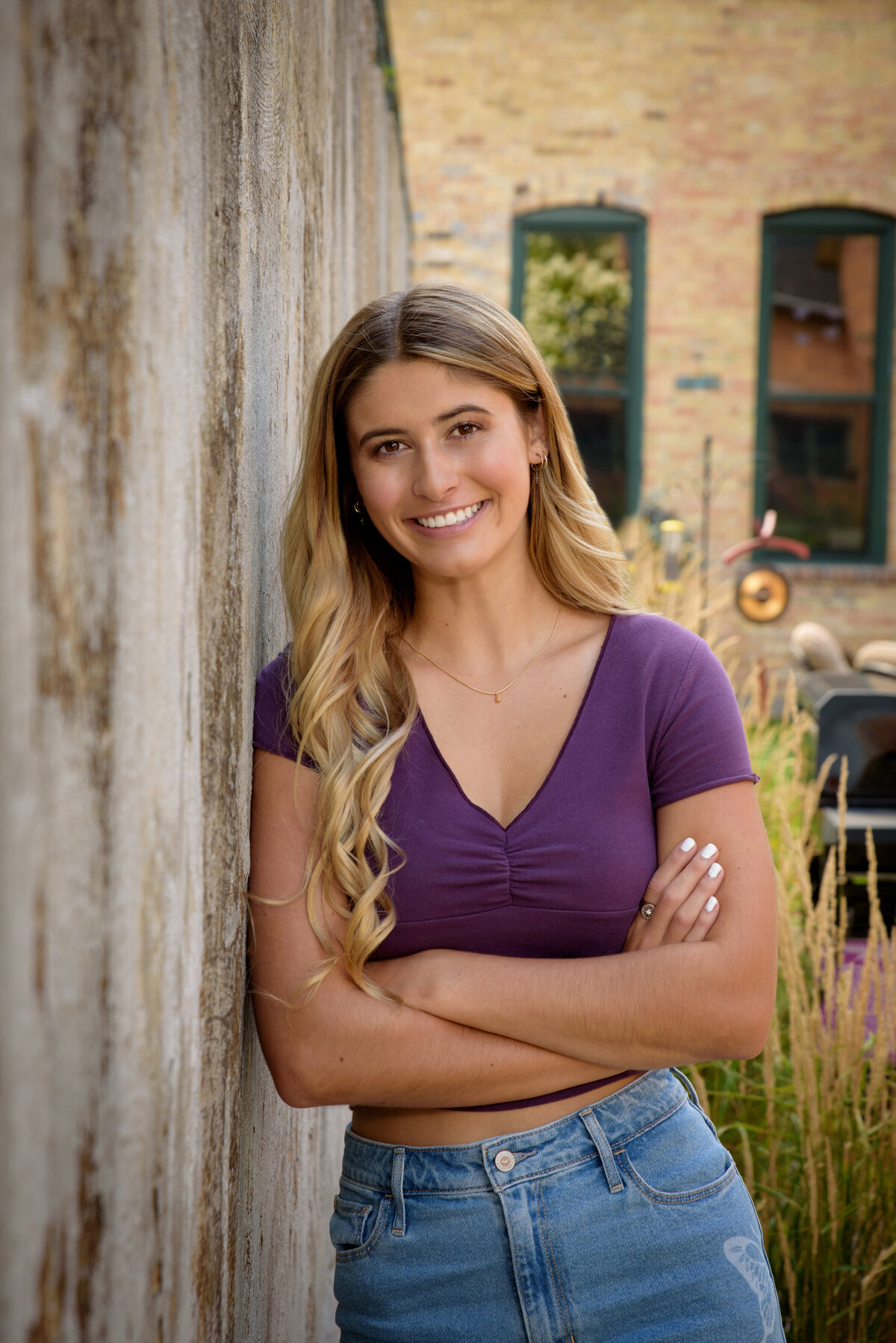 De Pere High School senior girl wearing a purple shirt and jeans aleaning against a cement wall by a brick building in an urban setting by Voyager Park in downtown De Pere, Wisconsin