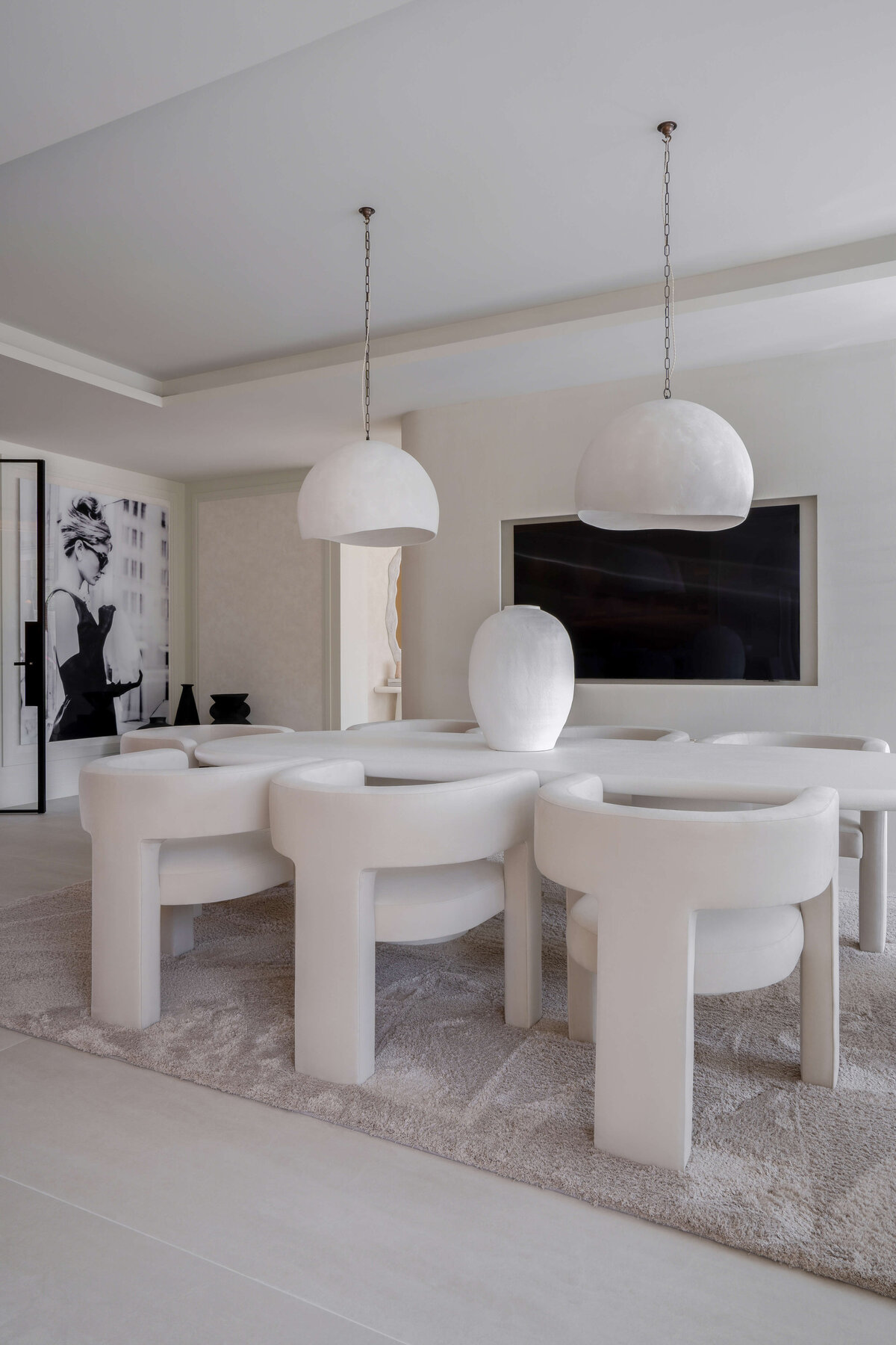 A modern dining room with a minimalist aesthetic, featuring a large oval table surrounded by six curved white chairs. Two large, white dome-shaped pendant lights hang above the table. The room is decorated in neutral tones with a plush, light-colored rug underneath the table and chairs. Behind the table, a large television recessed in the cream wall. is A black-and-white portrait is visible on the wall in the background, adding a touch of elegance to the space.