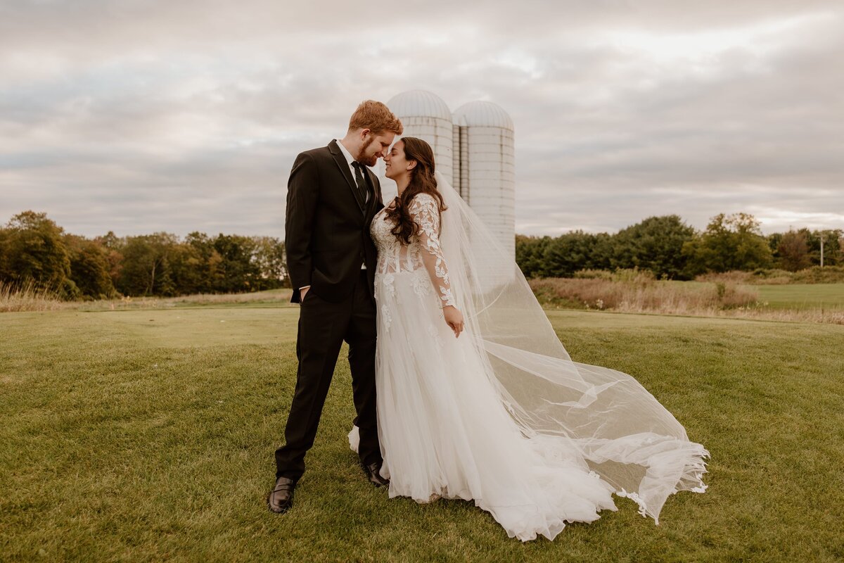A bride and groom share a tender moment at The Links at Union Vale, standing in front of two large silos under a cloudy sky. The bride wears a long-sleeved lace gown with a flowing veil, while the groom is dressed in a black suit. They gently lean into each other, creating a romantic and intimate scene amidst the rural, serene landscape.