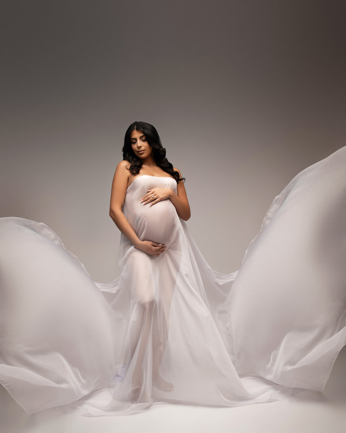 pregnant woman wrapped in silk flowing white fabric standing in front of white grey backdrop for studio maternity