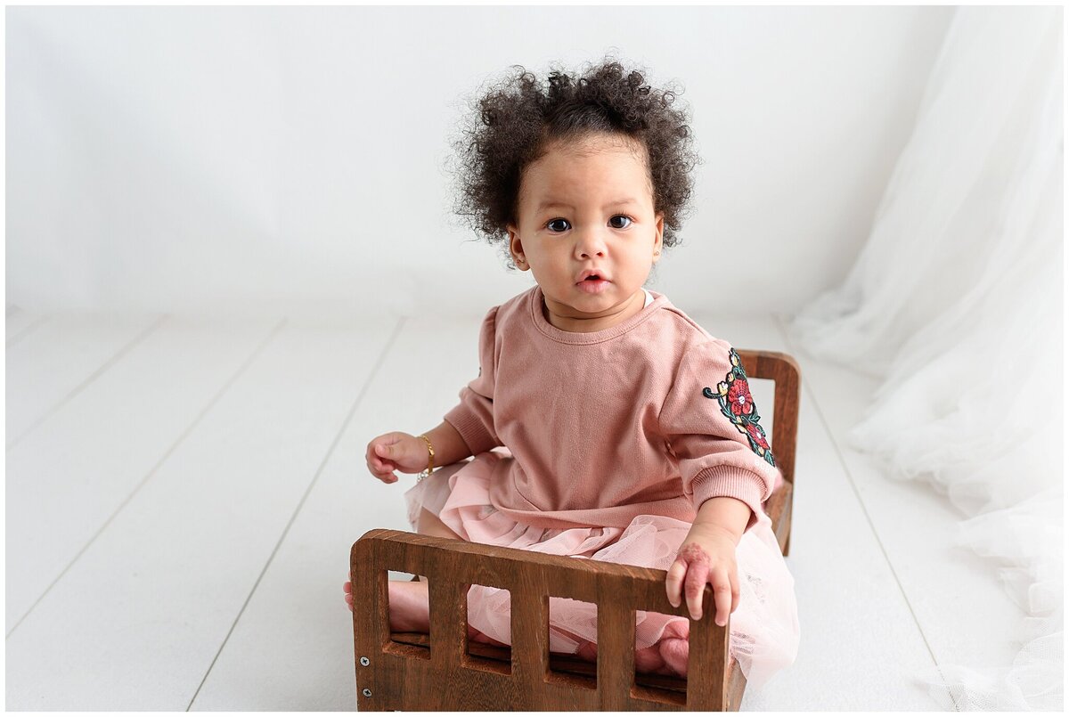 six month old photo session with baby girl sitting on a bed prop and looking at the camera.