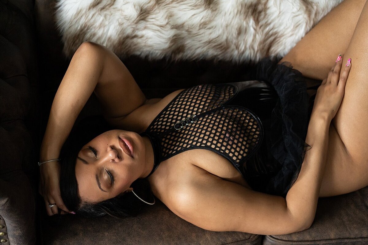 woman laying on her back on a leather couch, with eyes closed in a fishnet bodysuit.