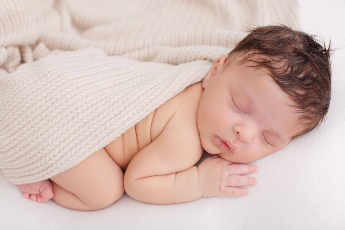 Newborn baby sleeping on belly with hand up by face and toes peeking out under a beige textured blanket - newborn photos in portland, Oregon