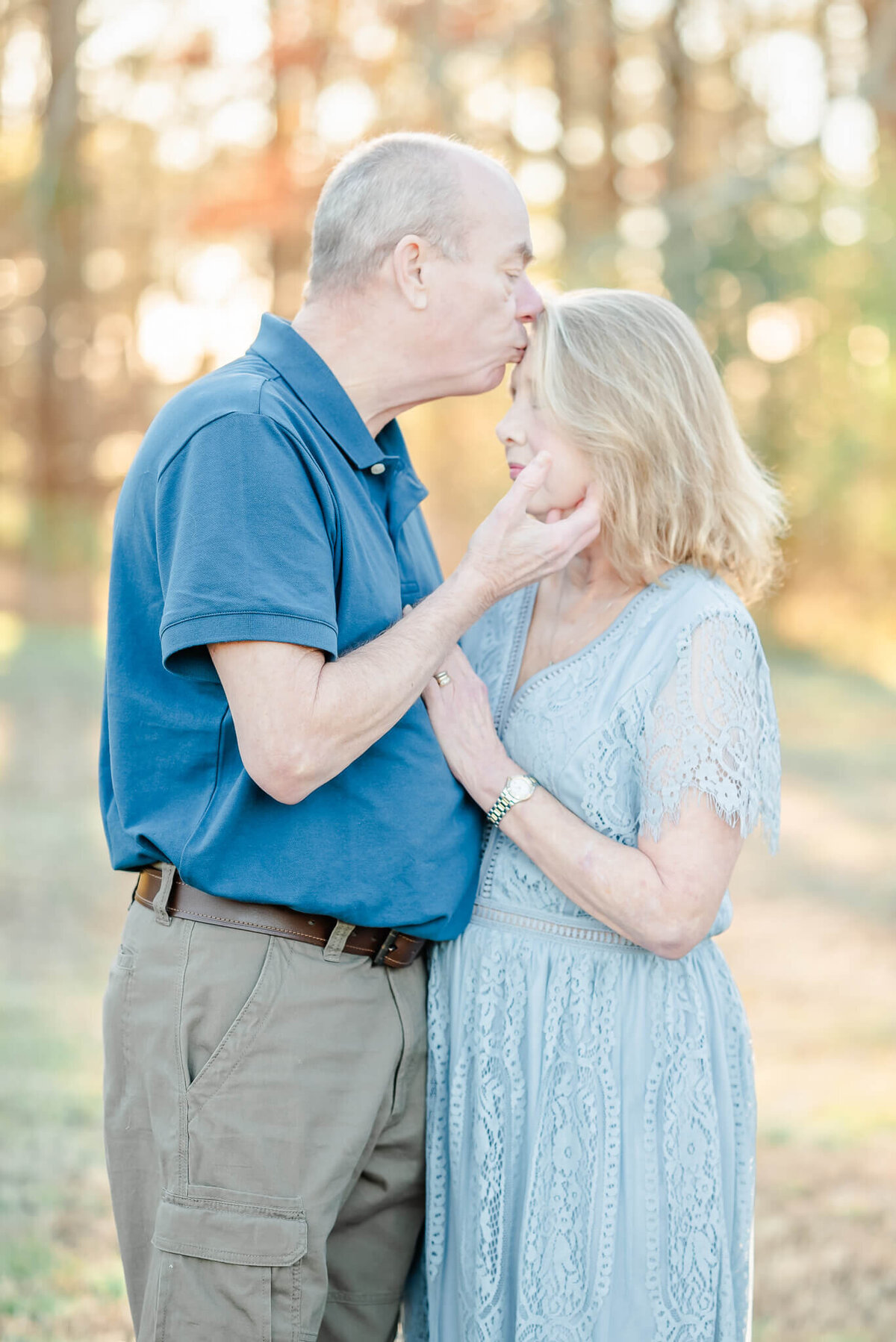 An older man, wearing a navy shirt, kisses his wife on the forehead.  His wife, wearing a blue lace dress, closes her eyes and puts her hand on his chest.
