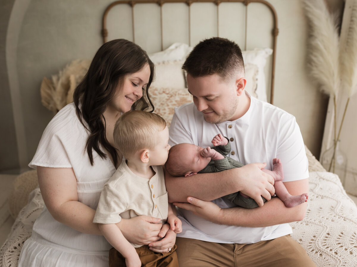 parents sitting on bed holding two year old son and newborn baby boy for studio portraits cleveland newborn photographer
