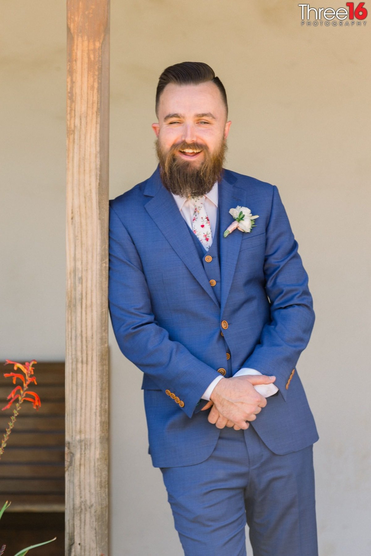 Groom leans up against a post as he poses for photos with a large smile on his face