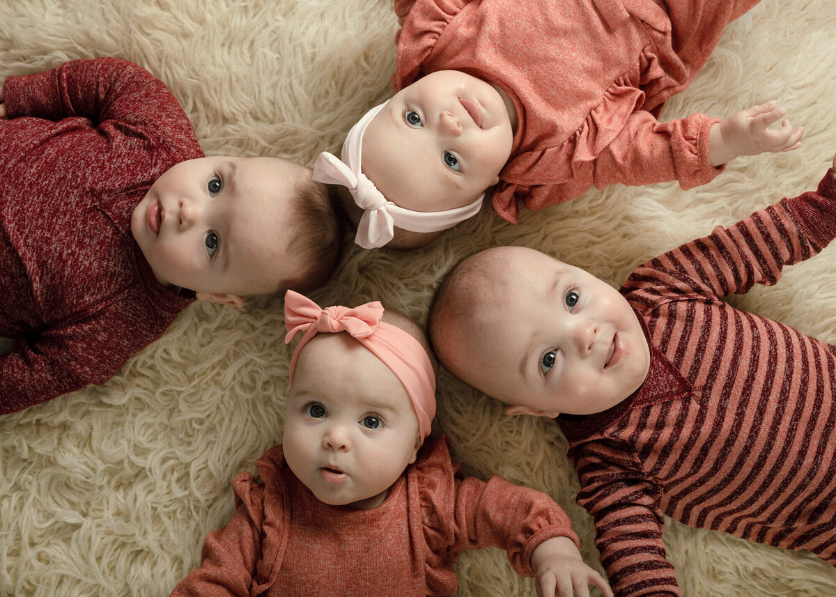 Quadruplets at 6 months old all laying on a creme colored rug looking up. All dressed in shades of coral.