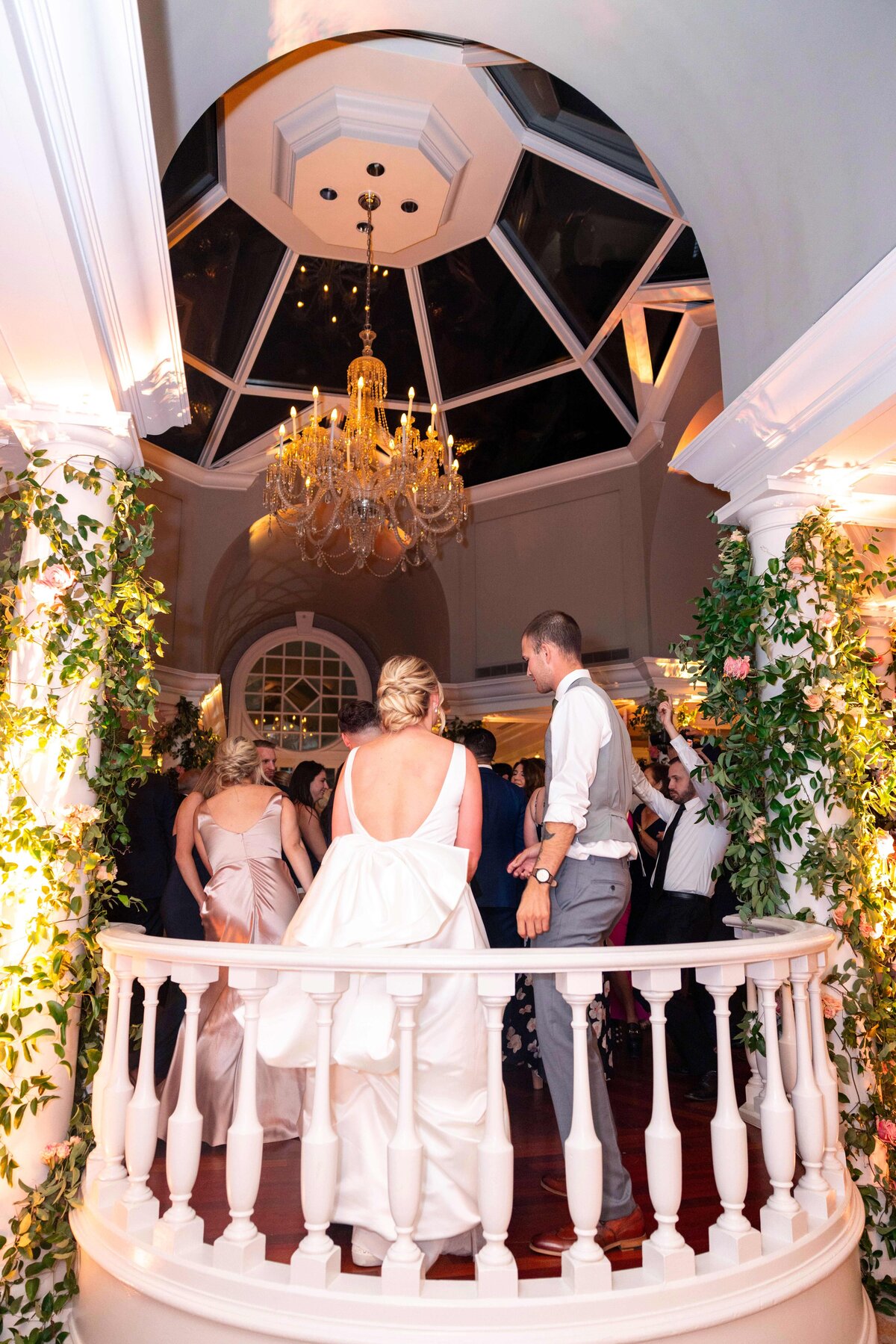 A bride and her guest stand together on a balcony overlooking a dance floor at a wedding reception. The venue has a large chandelier, glass ceiling, and is decorated with greenery. Guests in formal attire are dancing below.
