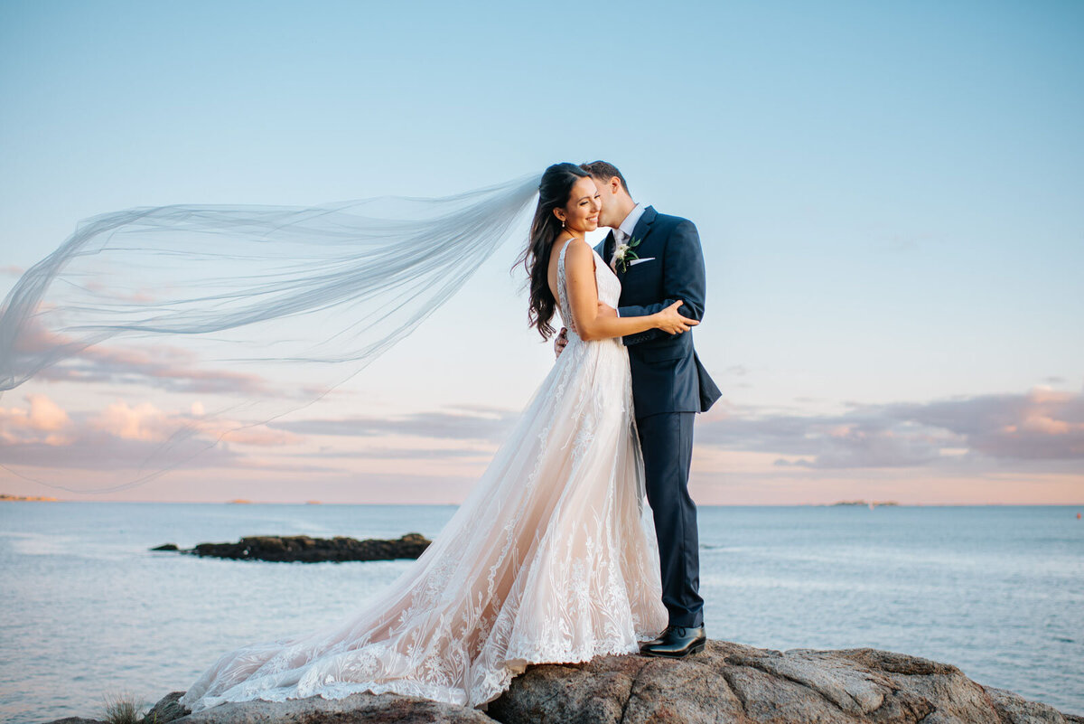 couple kissing on rocks veil blowing in the wind by the ocean at endicott college wedding