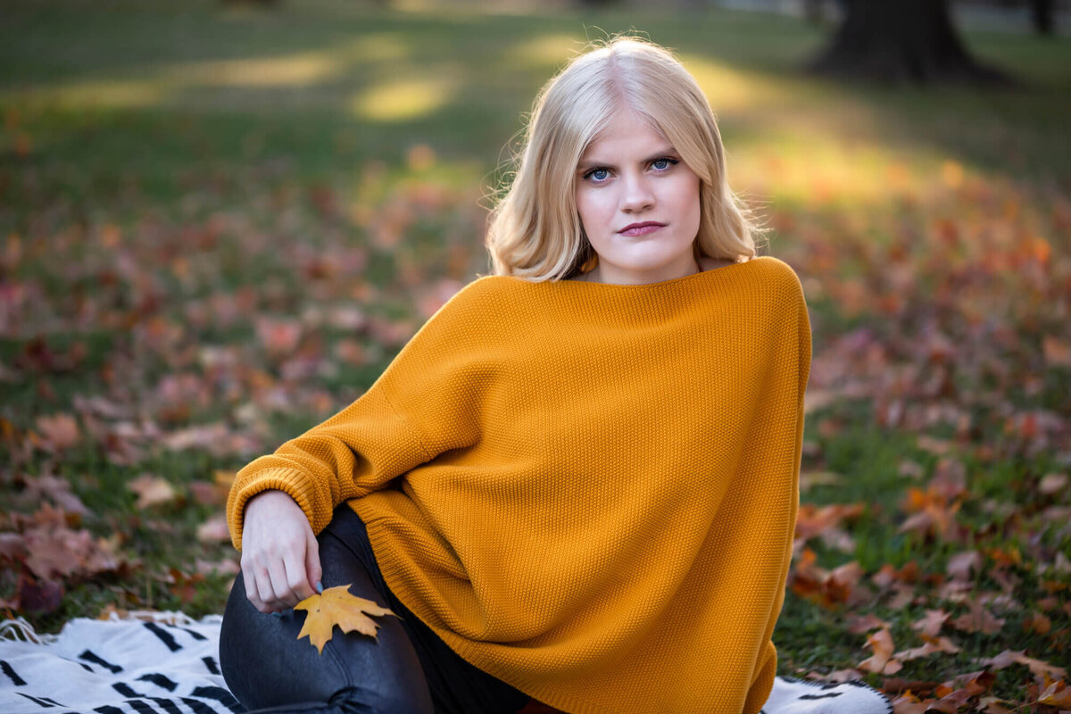 A beautiful blonde teen poses for a Fall senior portrait wearing a golden yellow sweater and holding a golden maple leaf sitting on a black and white blanket. Captured by Springfield, MO senior photographer Dynae Levingston.