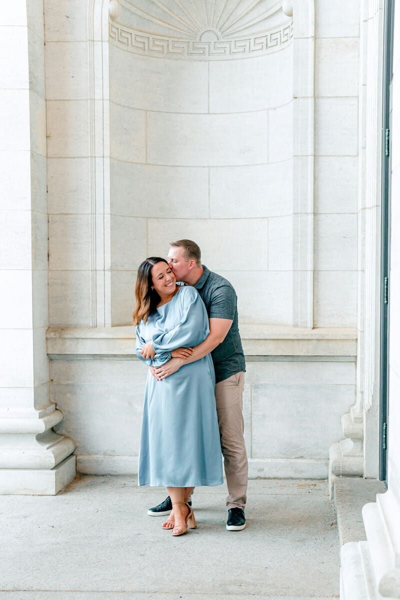 A couple sharing a loving embrace under a beautiful archway, captured during their engagement photo session. The woman is wearing a light blue dress, and the man is in a casual shirt and pants, kissing her cheek affectionately.
