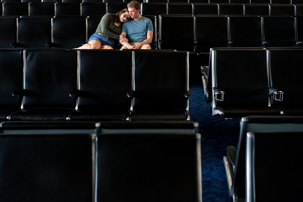 woman and man sit together while holding arms in airport seats near boarding gate