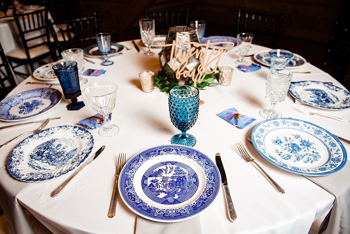 Wedding Reception table at Sycamore Farms set with a white table cloth, mismatched blue vintage china plates, golf flatware and mismatched blue vintage glass water goblets. Blue and white goo goo cluster wedding favors. Low floral centerpiece at the center of the table.