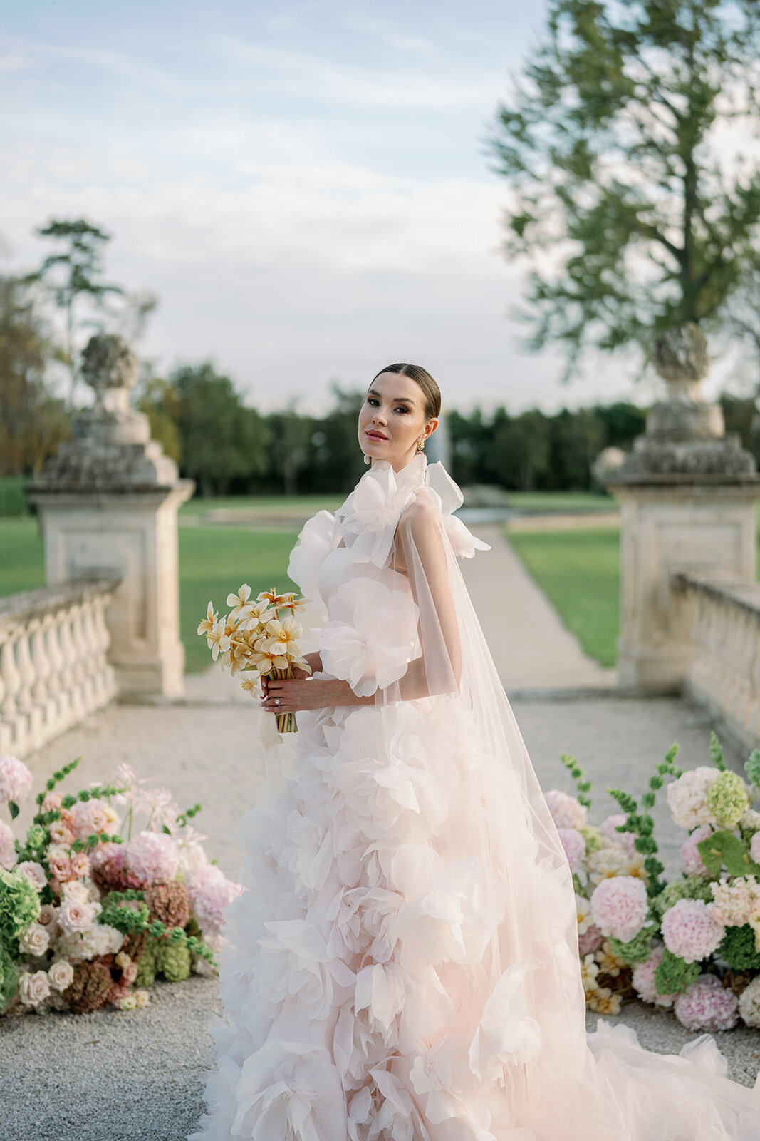 Bride posing beside grand floral installation on stone terrace with sweeping garden views at Château de Tourreau.