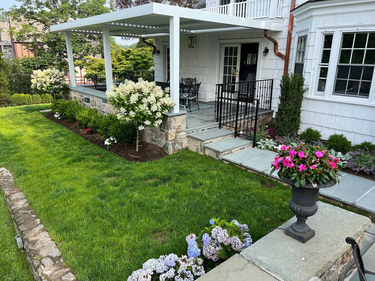 Elegant porch with a white pergola, black wrought iron railing, and lush landscaping featuring hydrangeas, vibrant flowers, and manicured greenery.
