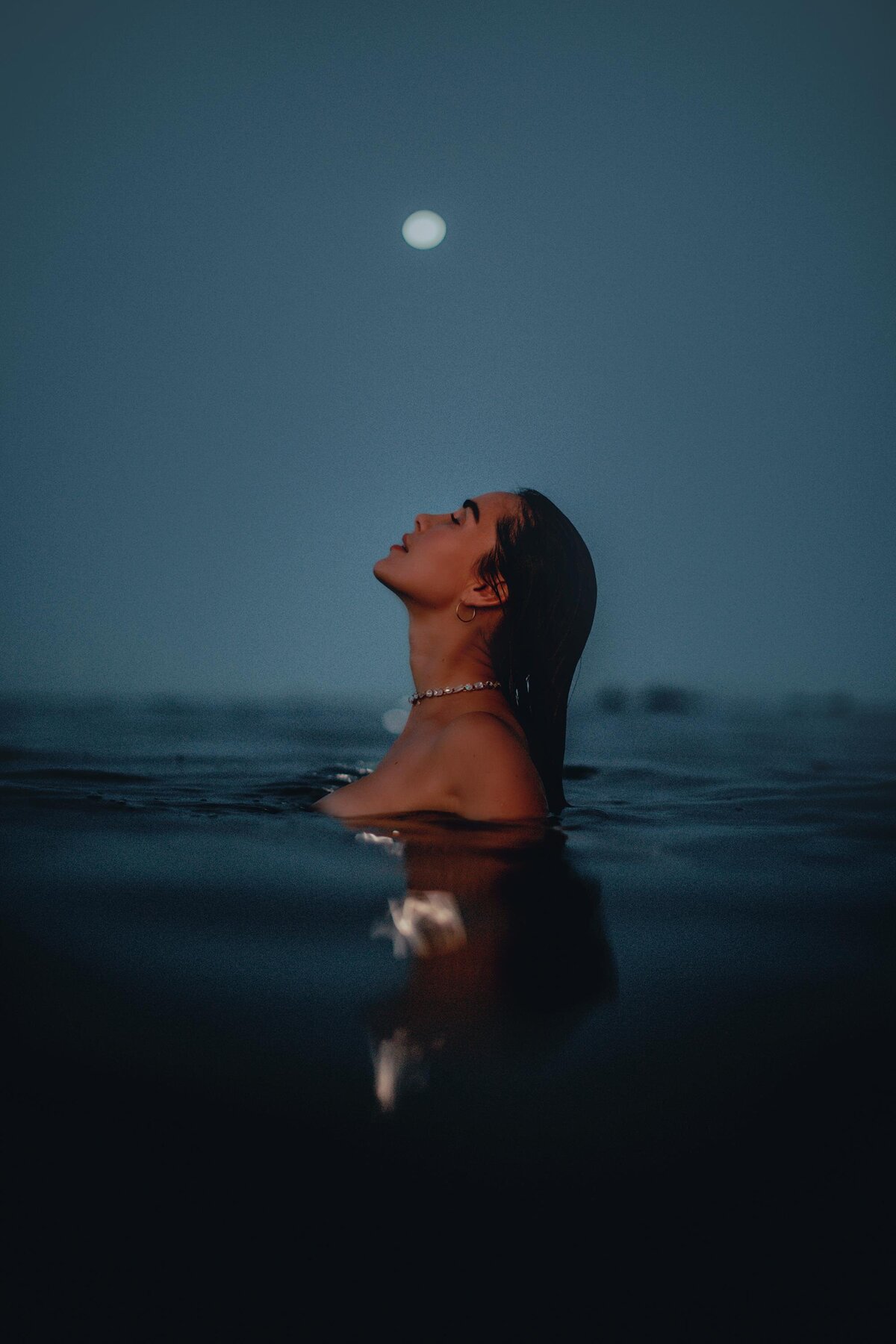 Stunning woman swims in the ocean with her head turned toward the moon during her boudoir session in Hawaii