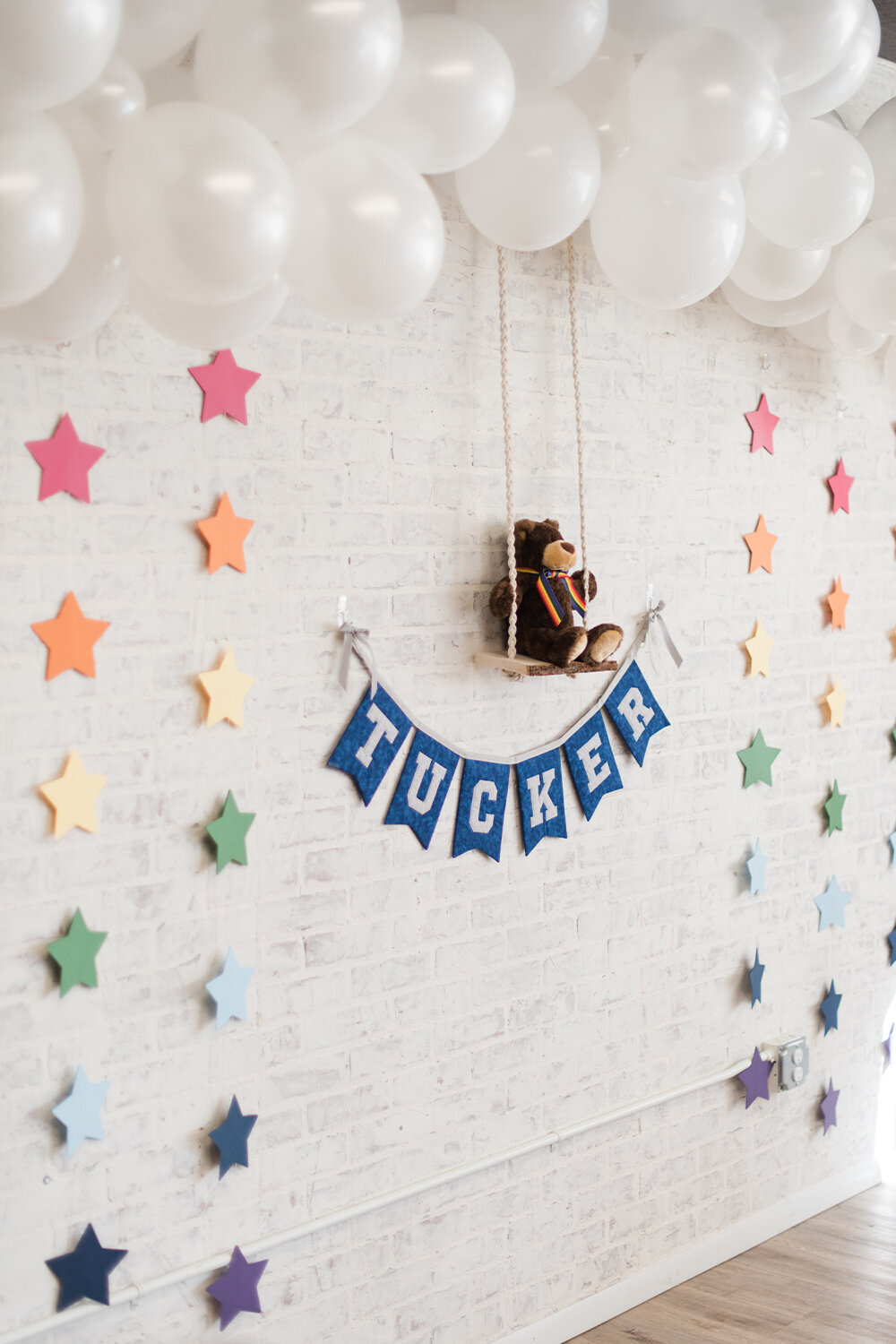 Woodland baby boy shower backdrop against a white brick wall featuring rainbow star garland, white balloon clouds, and a teddy bear sitting on a wooden swing with a fabric garland displaying the baby’s name.