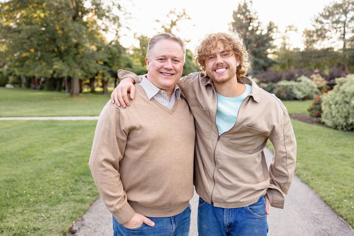 A father and son standing side by side, arms around each other, smiling warmly at the camera in a serene park.