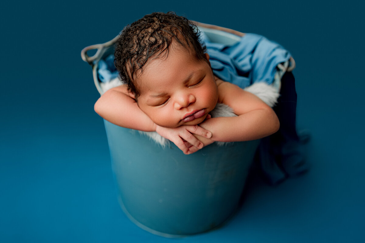 In a serene scene of Charlotte newborn photography, a tiny baby sleeps peacefully, arms folded on the rim of a blue bucket. The bucket is cozily lined with a soft blue blanket, perfectly complemented by the matching backdrop.