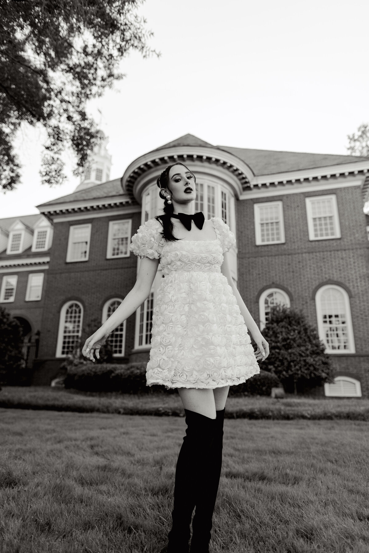 Dramatic black and white editorial portrait of a woman in a textured floral mini dress with a black bow and over-the-knee boots, posed in front of a historic brick mansion in Greenville, South Carolina. Captured by Dani Lacey Photographs.