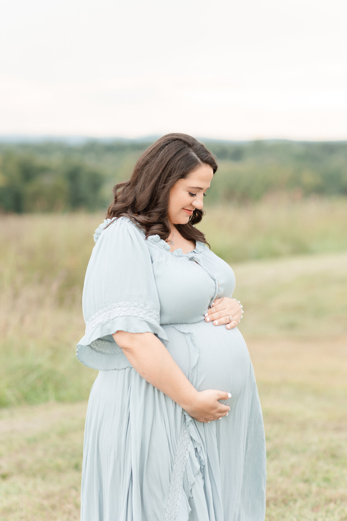 A mother holding her belly outside in a field wearing a blue dress at a DC Newborn Photography session