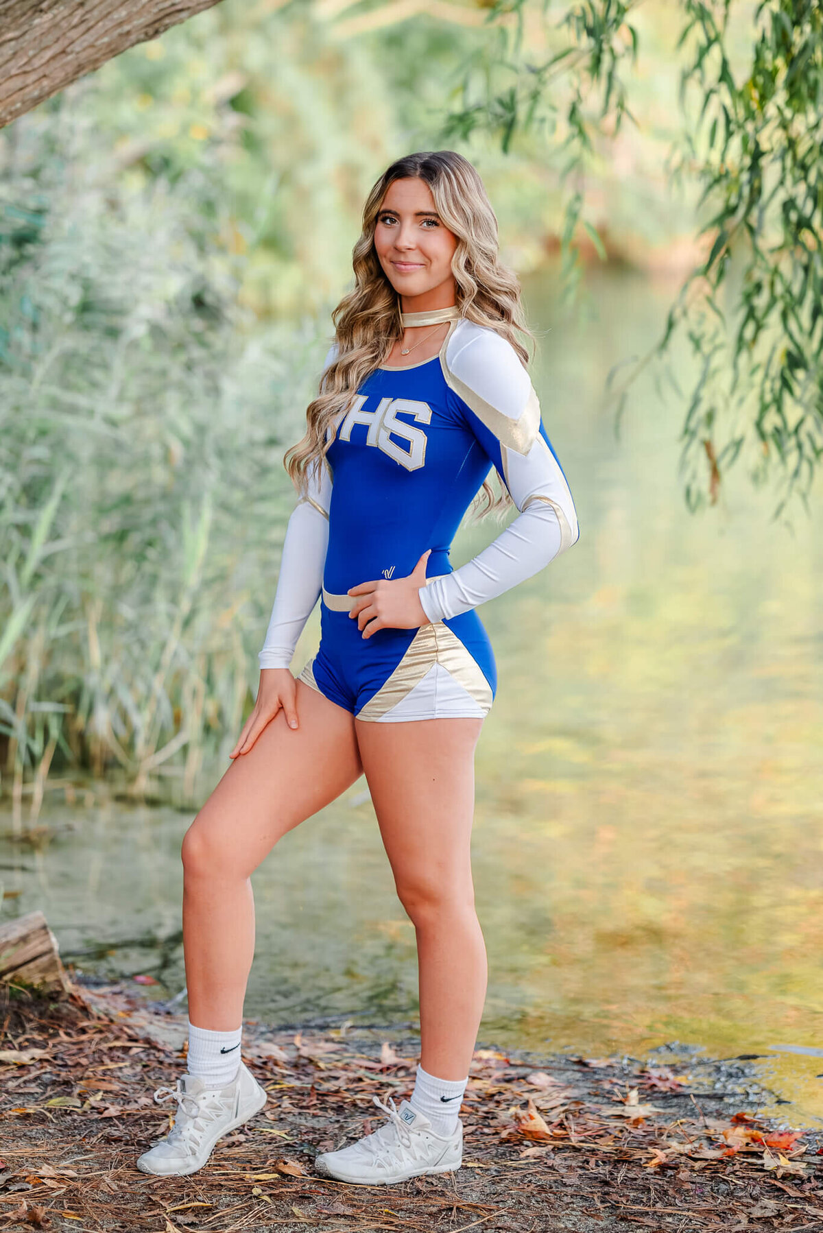 A high school senior, wearing her blue, white and gold cheer uniform, stands under a willow by the lake at Oak Grove Lake Park in Chesapeake, VA.