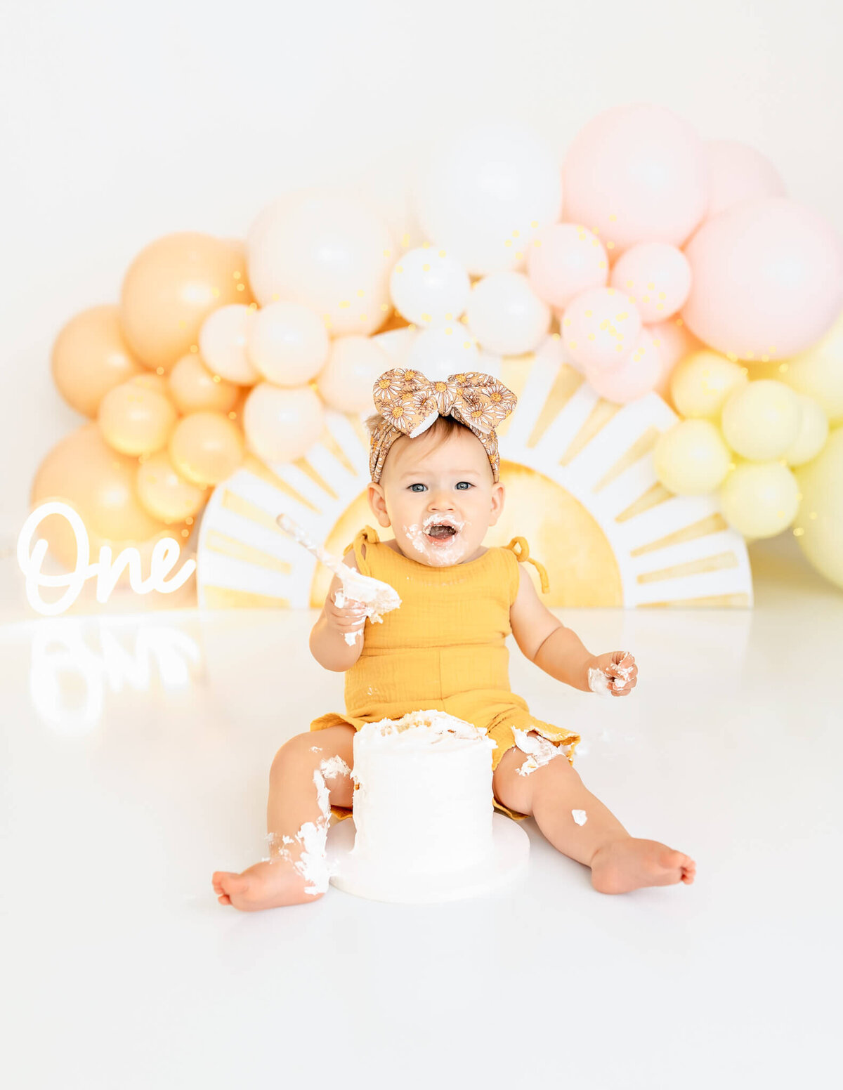 A baby wearing a yellow outfit and a bow sits on the floor surrounded by pastel balloons. The baby has a cake in front, with frosting on their face and hands. A lit "One" sign is visible, indicating a first birthday celebration.