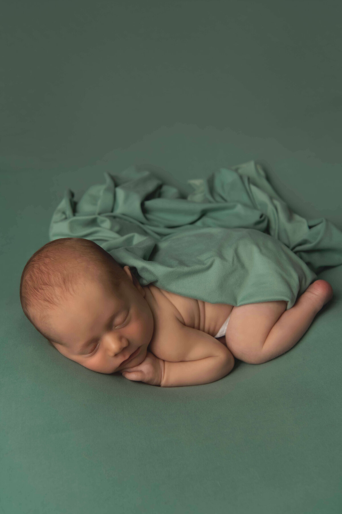 A newborn boy posed carefully in the bum up pose on a sage green backdrop. You can see all of his newborn details, including his back fat rolls. He has a sage green blanket covering him, as it flows into the backdrop to create interest.