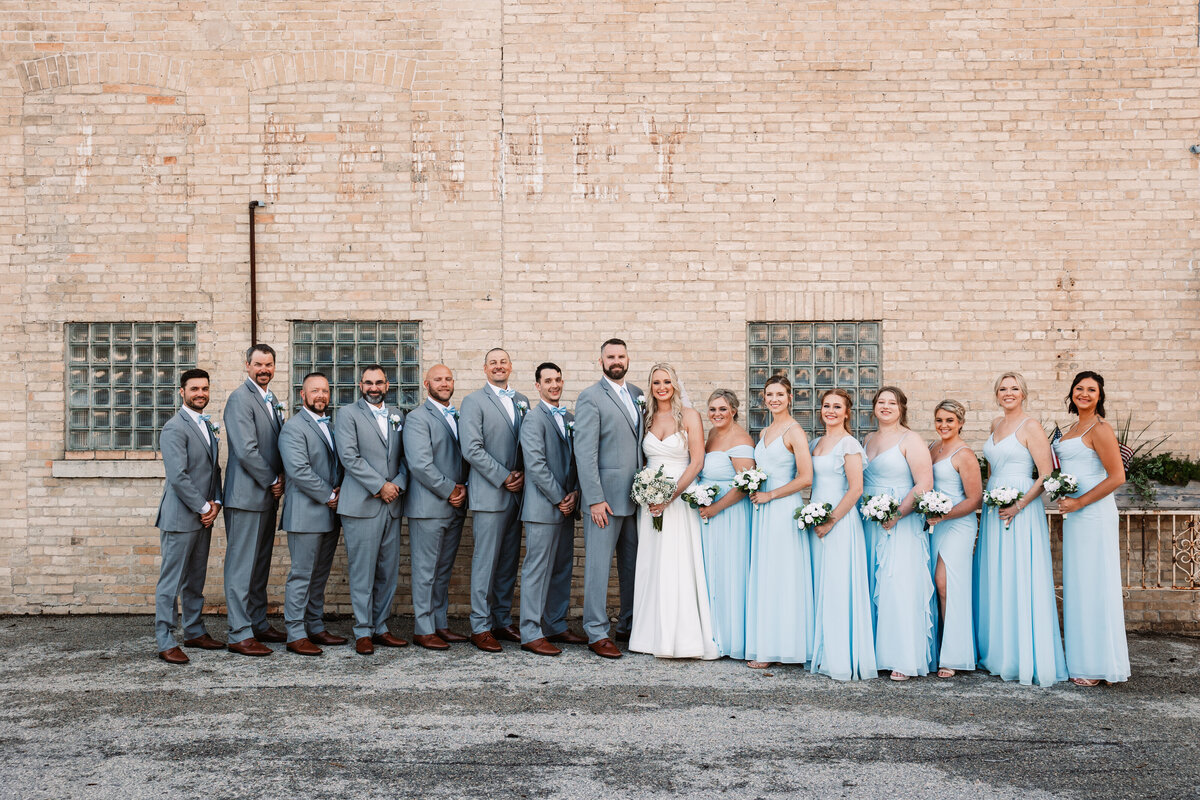 Newlyweds stand with their wedding party against a light brick wall