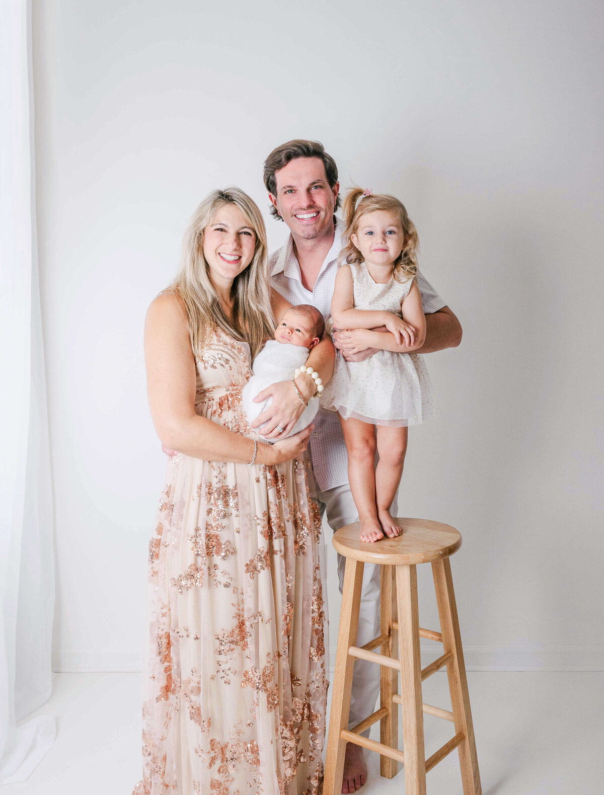 A blond haired mom dressed in a sparkling gold gown holding her newborn baby with her husband and young  daughter posing next to a window in a white room.