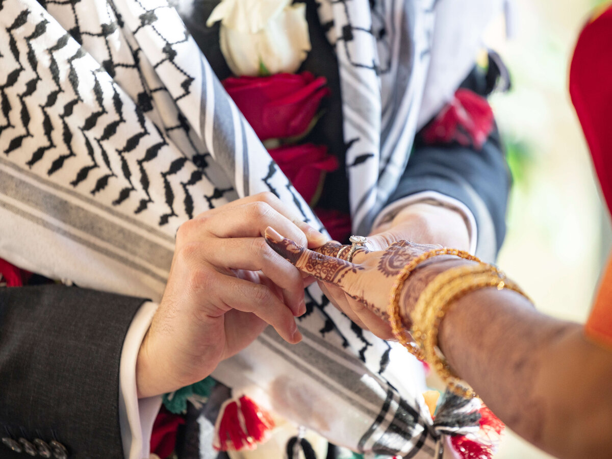 A close-up of two people exchanging rings. One hand has intricate henna designs and gold bangles, while the other wears a dark suit. A patterned shawl with red and white flowers is visible in the background.