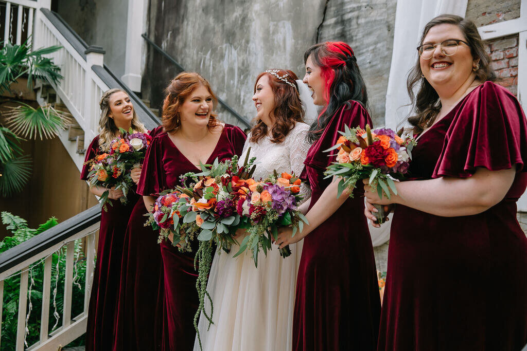 A bride in a white dress stands with five bridesmaids in matching burgundy dresses, all holding bouquets, on wooden stairs decorated with pumpkins and greenery.