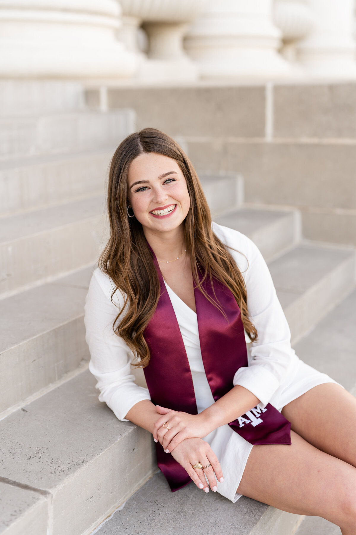 Texas A&M senior girl sitting on stairs and leaning on step while wearing white dress and Aggie stole and smiling at the Administration Building