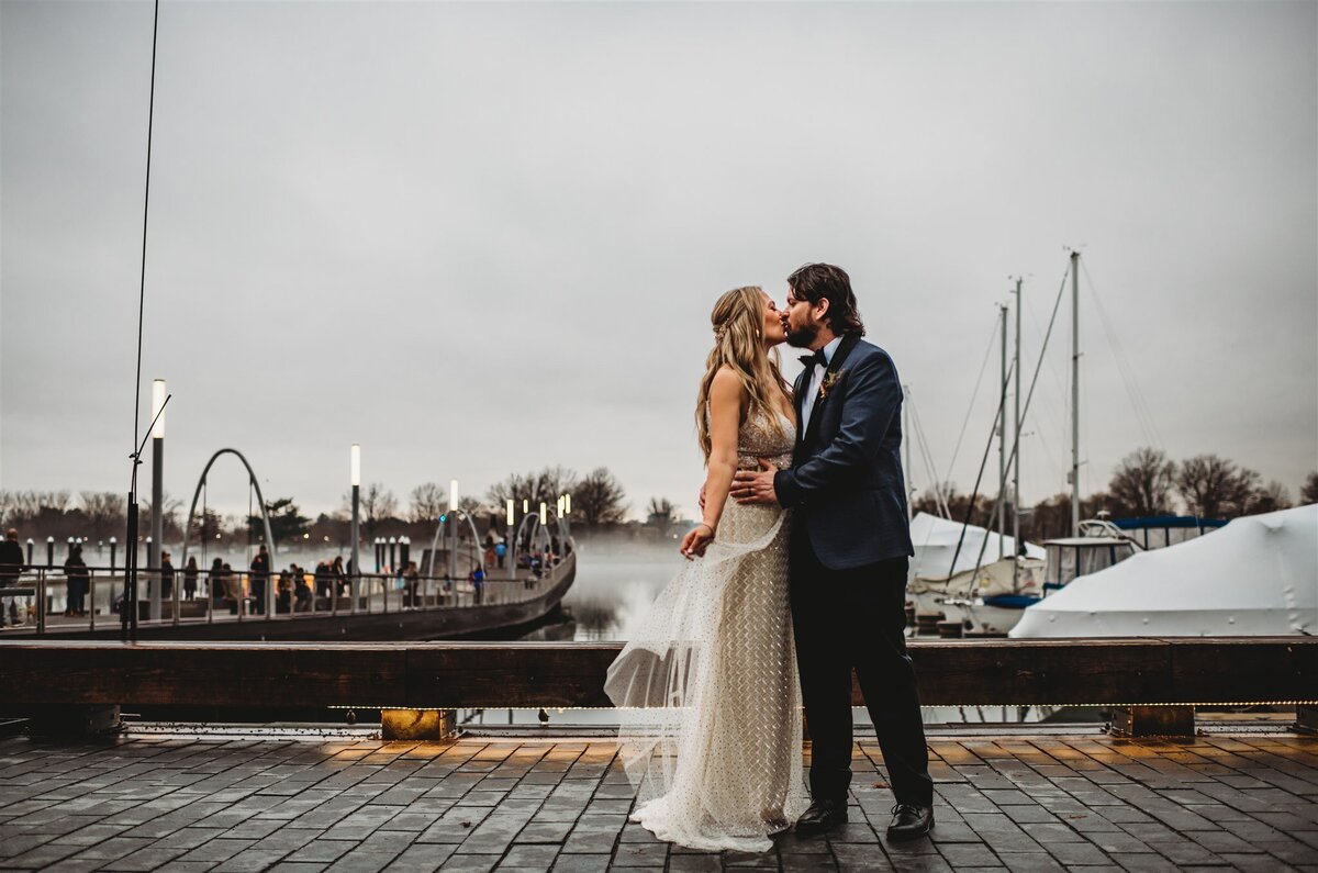 Bride and groom standing on a stone pier next to parked boats out of dock and kiss one another as the wind blows the bride gown captured by Maryland wedding photographer