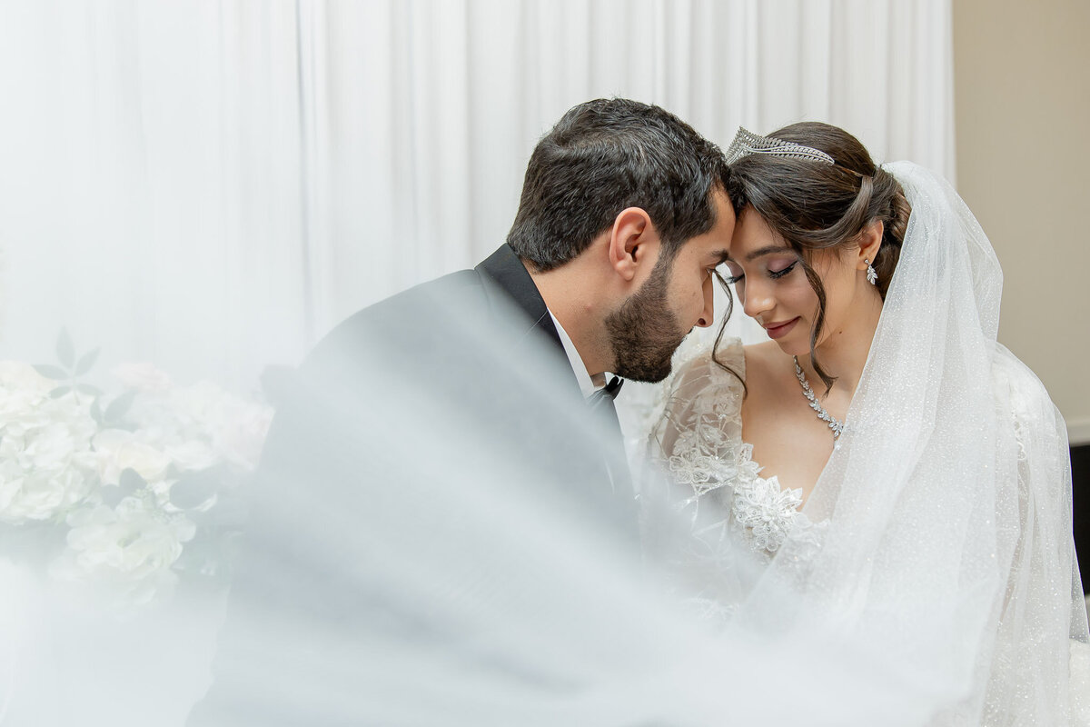 A bride and groom gently touch foreheads as they're sitting down. The brides veil sweeps across the room, slightly in front of the camera lens.