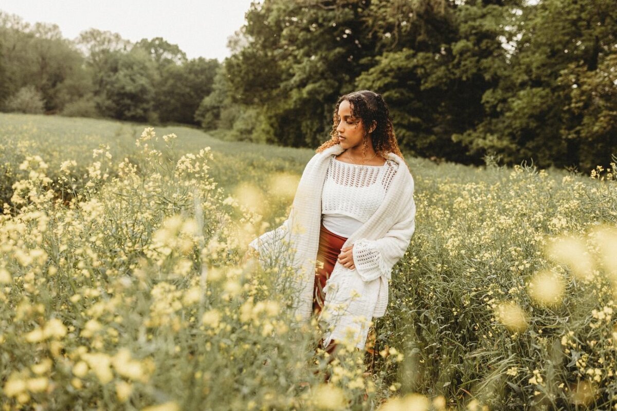 A woman with curly hair stands in a field of yellow flowers, wearing a white top and shawl, looking left.