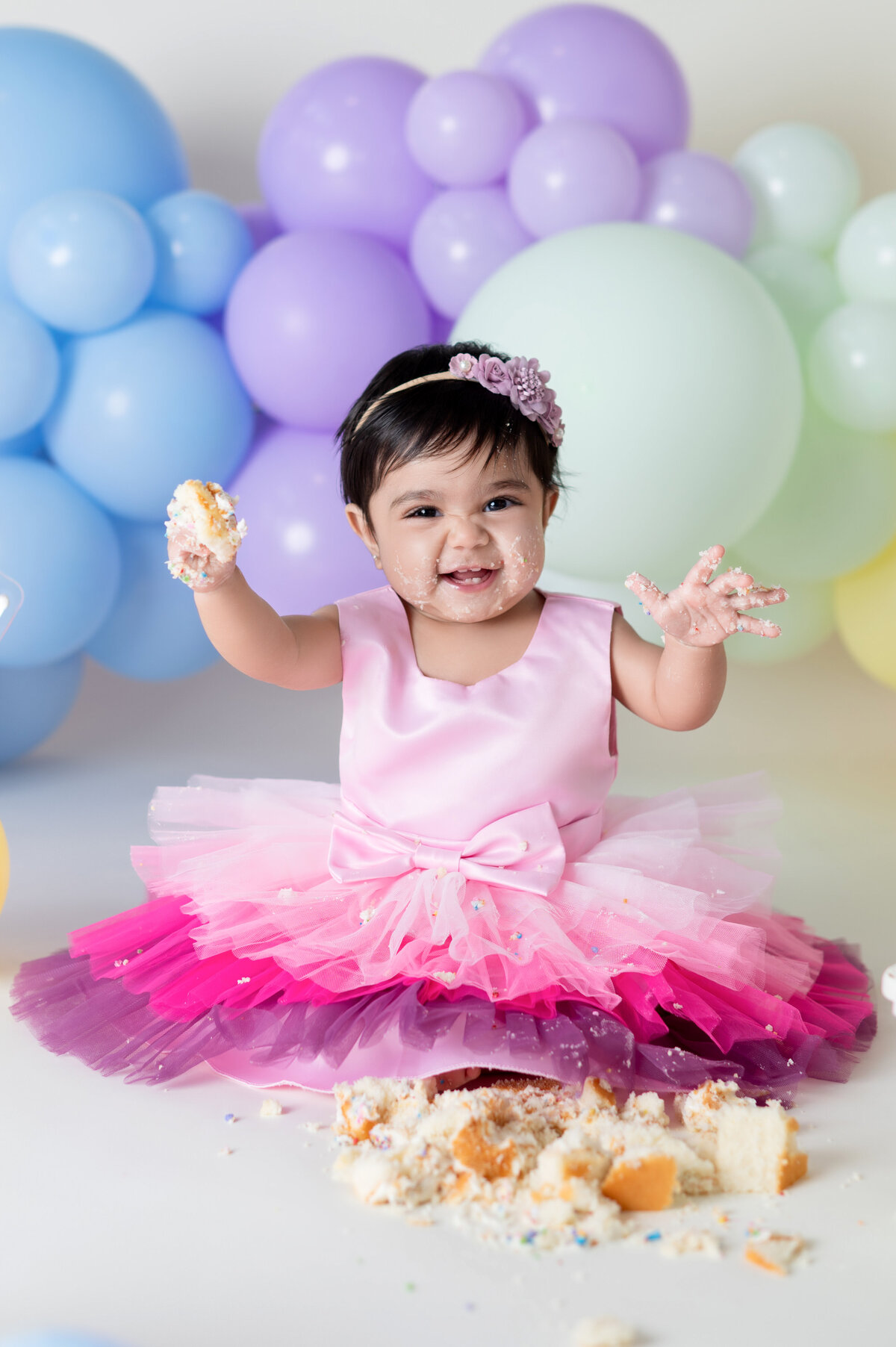 Smiling baby girl in a pink tutu dress enjoying her cake smash session with pastel balloon decor by Houston photographer Lissette G