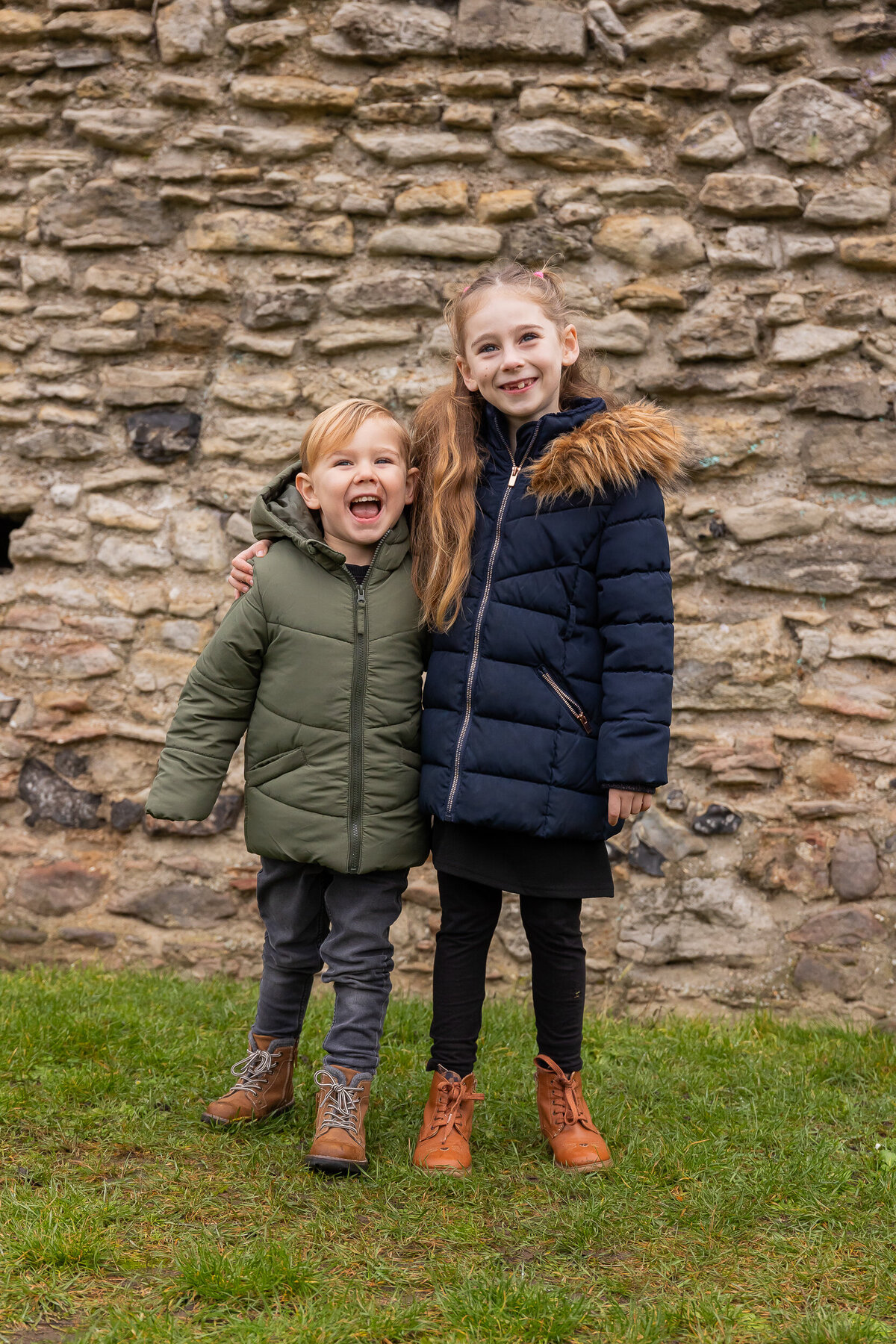 Two young children, a boy and a girl, standing and smiling in front of a stone wall. the boy is laughing and the girl is smiling with her arm around him.